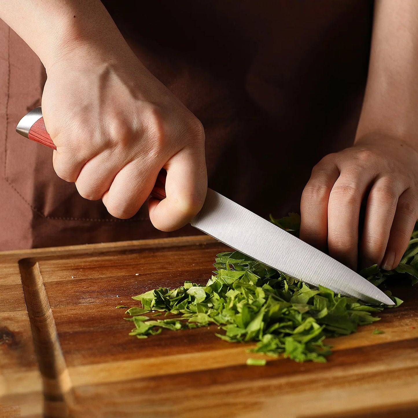 Person chopping green herbs on a wooden cutting board with a knife.