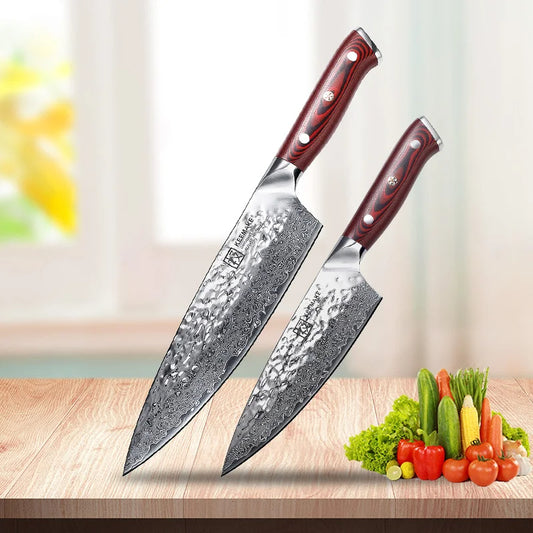 Two chef's knives with red handles on a wooden surface with vegetables in the background.